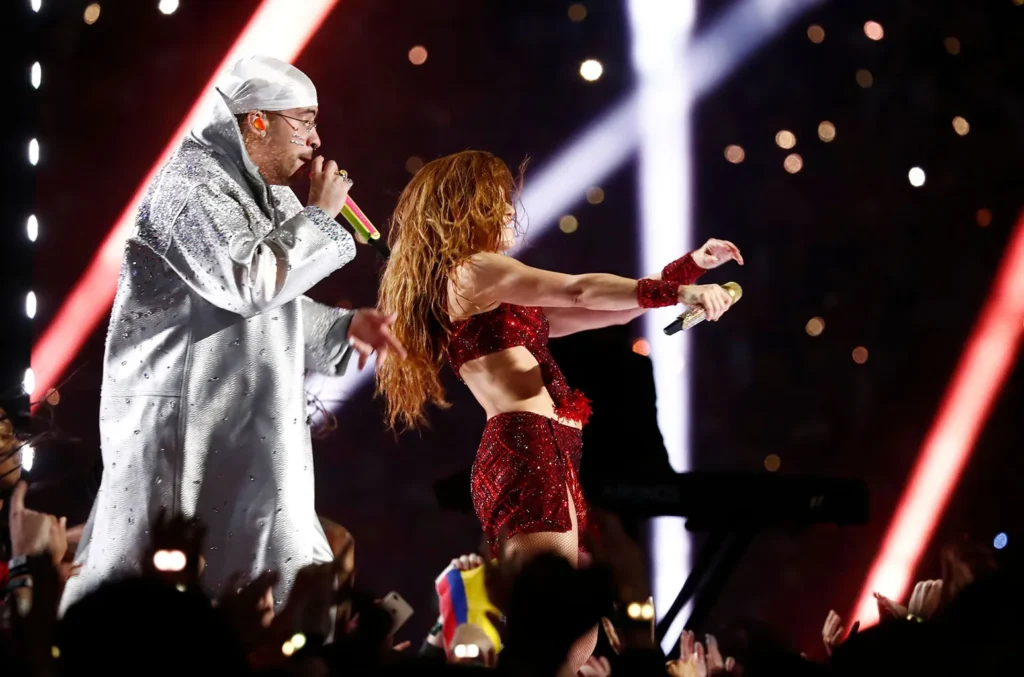 Bad Bunny and Shakira performing together during the 2020 Super Bowl halftime show, with Bad Bunny in a silver embellished hoodie and Shakira in a red sequined outfit dancing energetically on stage