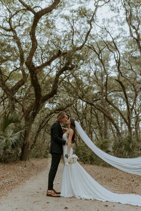 Bride and groom kissing under oak trees with flowing wedding veil at outdoor ceremony in South Carolina