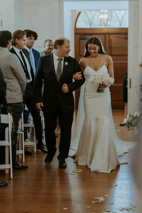 Bride walking down aisle with father in elegant mermaid wedding dress holding white rose bouquet