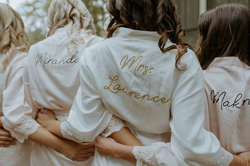 Bridesmaids in matching robes with embroidered names including Mrs. Lawrence before wedding ceremony