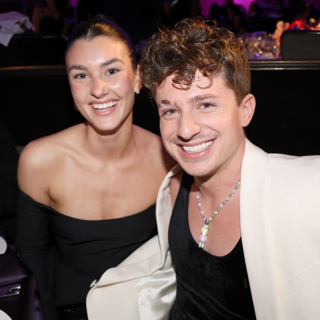 Charlie Puth and Brooke Sansone smiling together at entertainment industry event, Puth wearing cream blazer with pendant necklace