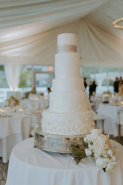 Four-tier white wedding cake with pearl details on silver stand at elegant tented reception