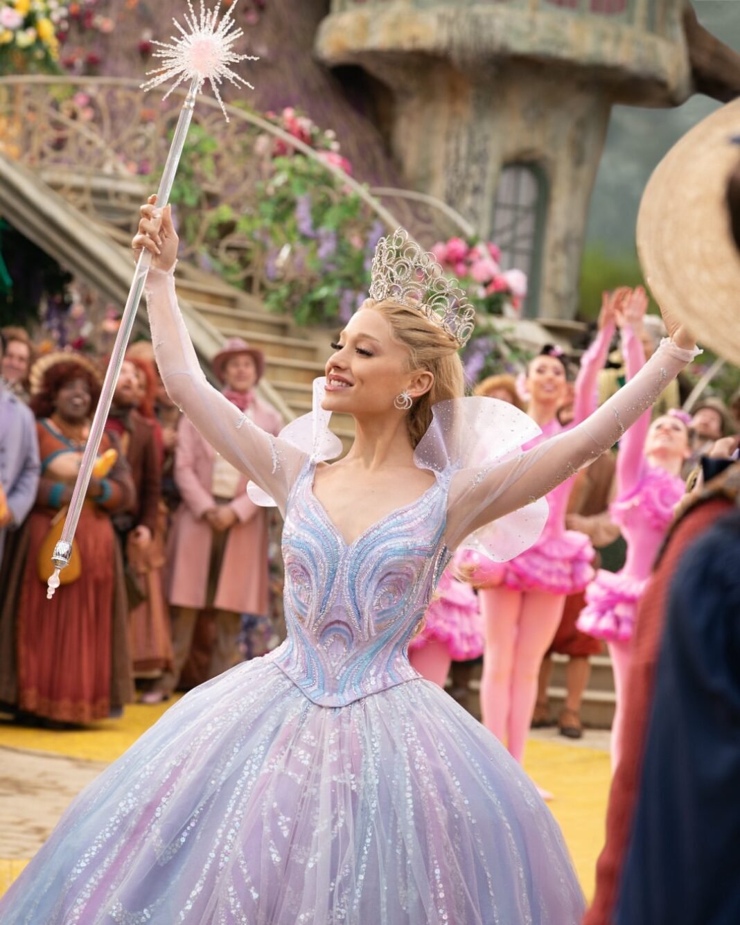 Glinda the Good Witch in an elaborate pale blue ball gown and sparkling tiara holds a glowing wand aloft during a celebratory scene in Wicked, surrounded by colorful Munchkinland citizens in a vibrant theatrical setting