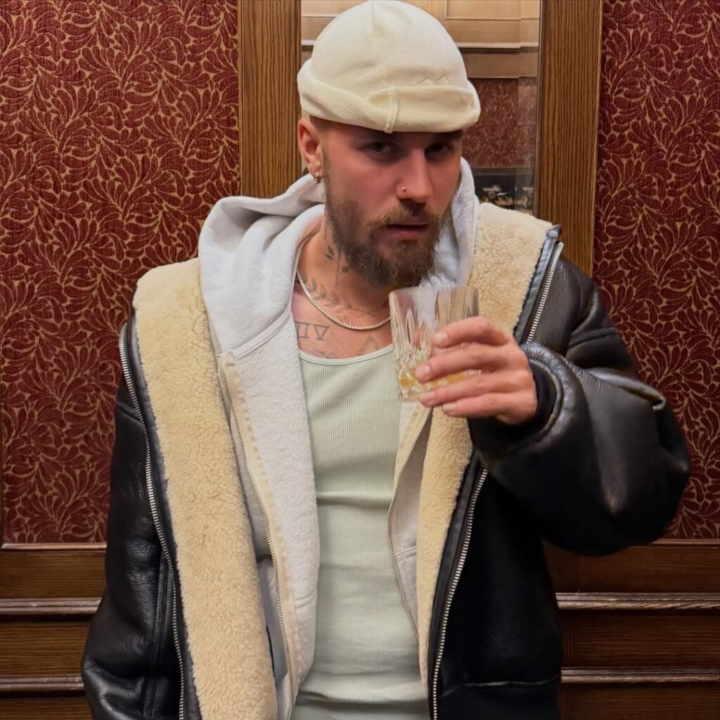 Justin Bieber holds champagne glass in elegant hotel interior wearing shearling jacket and cream cap