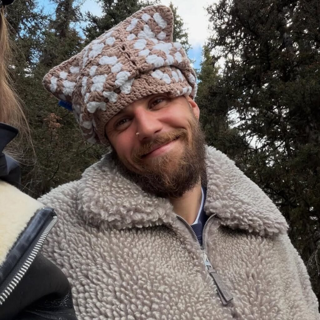 Justin Bieber smiles in selfie wearing whimsical knit mushroom hat and cozy shearling jacket in mountain forest