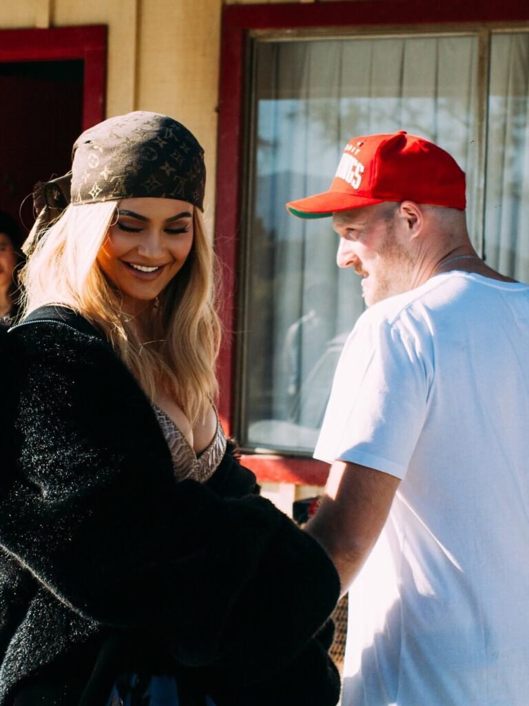 Kylie Jenner with blonde hair and black cap smiling with director on music video set