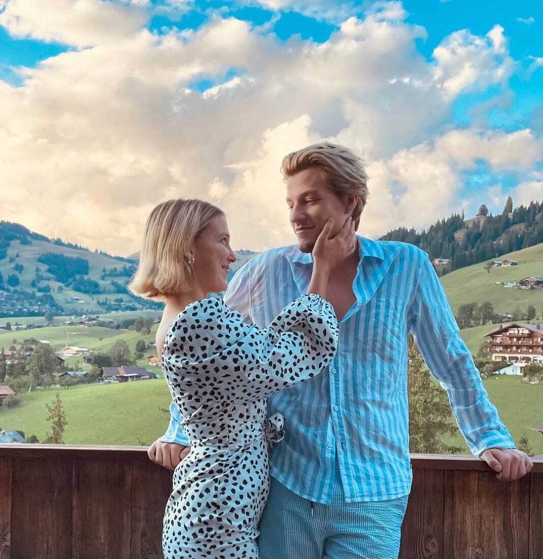 Millie Bobby Brown and Jake Bongiovi standing together on a wooden balcony overlooking a scenic Alpine valley with rolling green hills, traditional chalets, and dramatic cloudy sky in the background