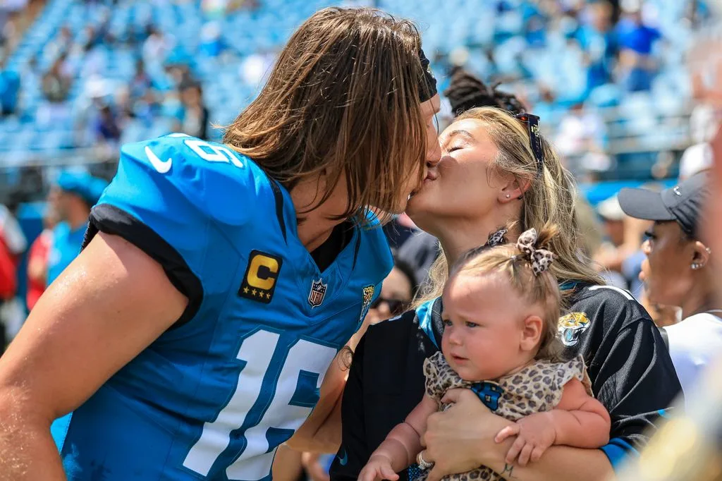 Trevor Lawrence in Jacksonville Jaguars blue jersey number 16 kissing wife Marissa while holding daughter Shae Lynn on field after game.jpg