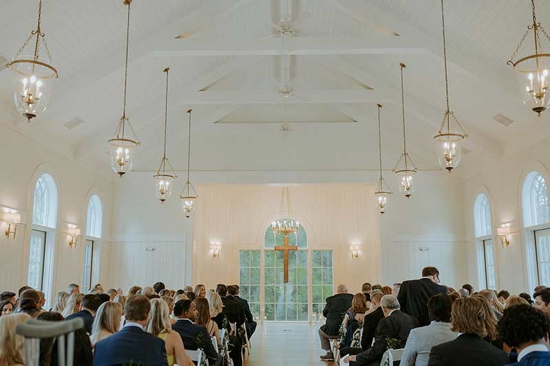 Wedding ceremony inside white chapel with hanging lanterns, arched windows and guests seated in wooden pews