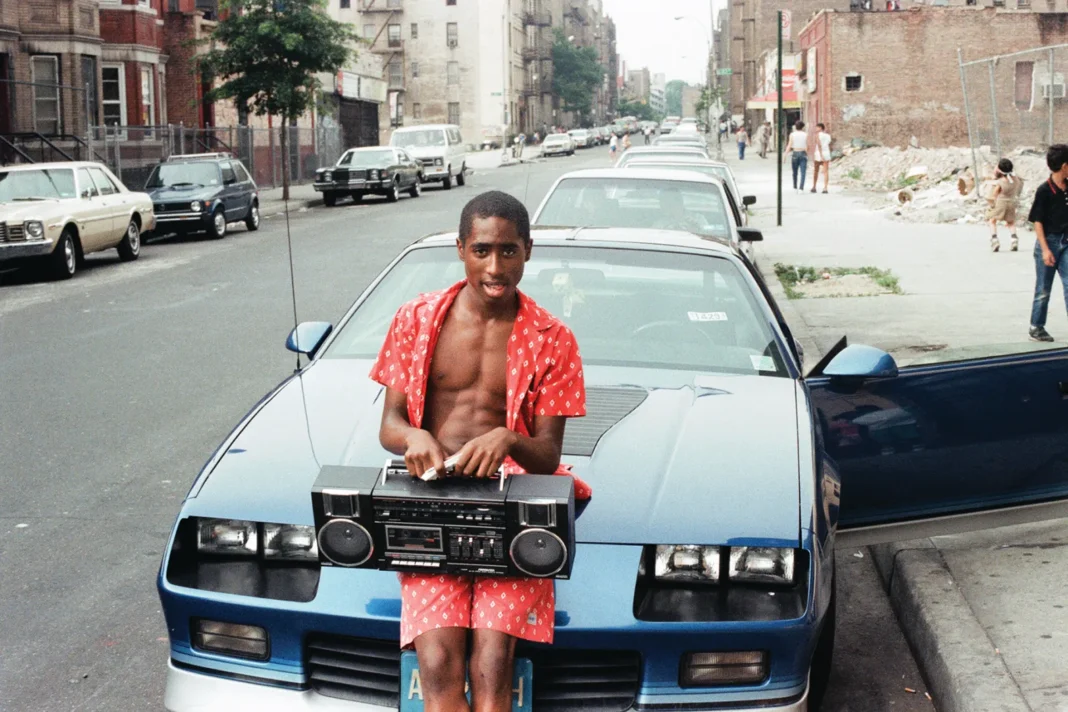 Young Black man in red shirt holds boombox in front of blue 1980s Camaro on urban street with brick row houses in background
