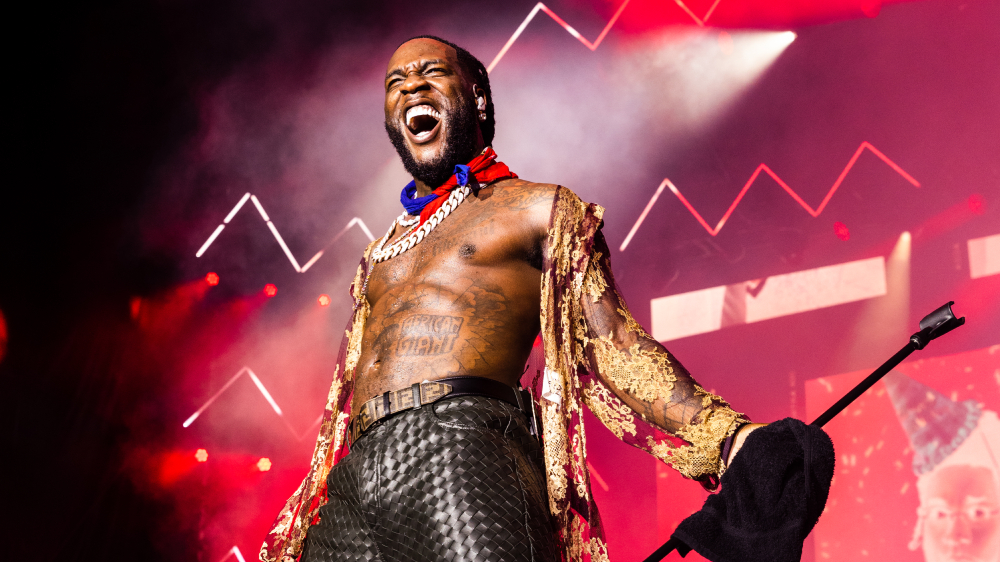 Burna Boy performing shirtless on stage with dramatic red and pink lighting, wearing a golden sequined jacket, chains, and sunglasses while singing into a microphone