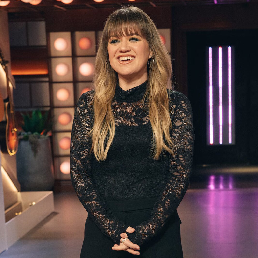 Kelly Clarkson smiles on the set of The Kelly Clarkson Show wearing a black lace long-sleeved dress with studio lighting visible in the background