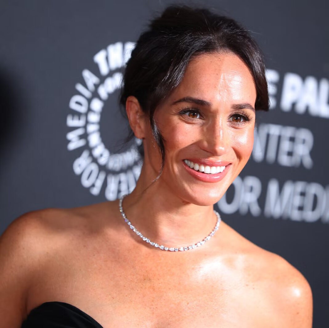 Meghan Markle wearing an elegant black strapless gown and diamond necklace at a media event, smiling at the camera on the red carpet
