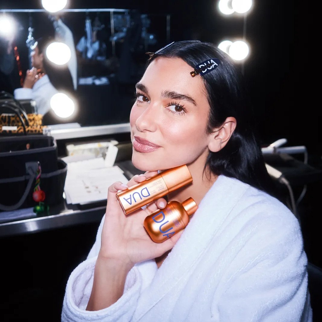 Model with DUA Skincare branded hair clip holding bronze cleanser and serum in vanity mirror setting, showcasing complete three-step routine from Dua Lipa and Augustinus Bader collaboration