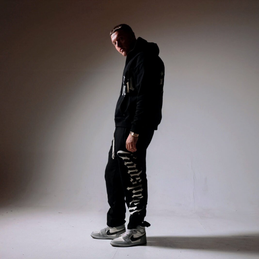 Toxic Rob stands in dramatic studio lighting wearing a black hoodie and sweatpants with 'Toxic Rob' text, paired with Nike sneakers