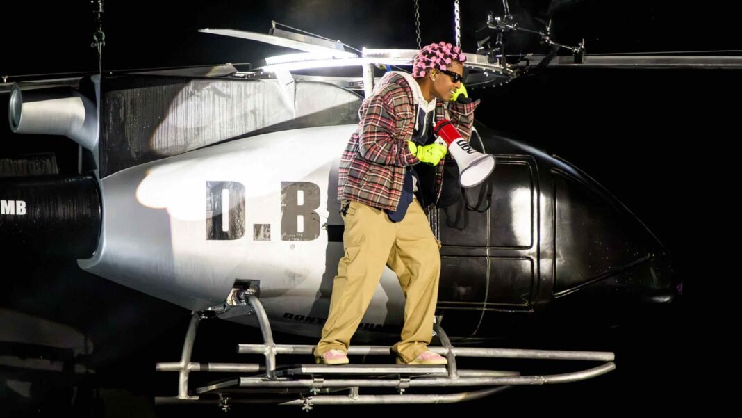 A$AP Rocky performing on stage standing on a helicopter prop with 'D.B' lettering, wearing a plaid jacket, pink beanie, and yellow pants