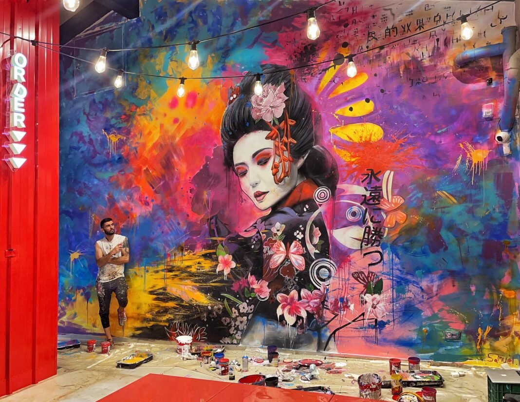 Street artist Samuel Hagai standing beside his large-scale mural featuring a geisha with vibrant colors, flowers, and Asian calligraphy