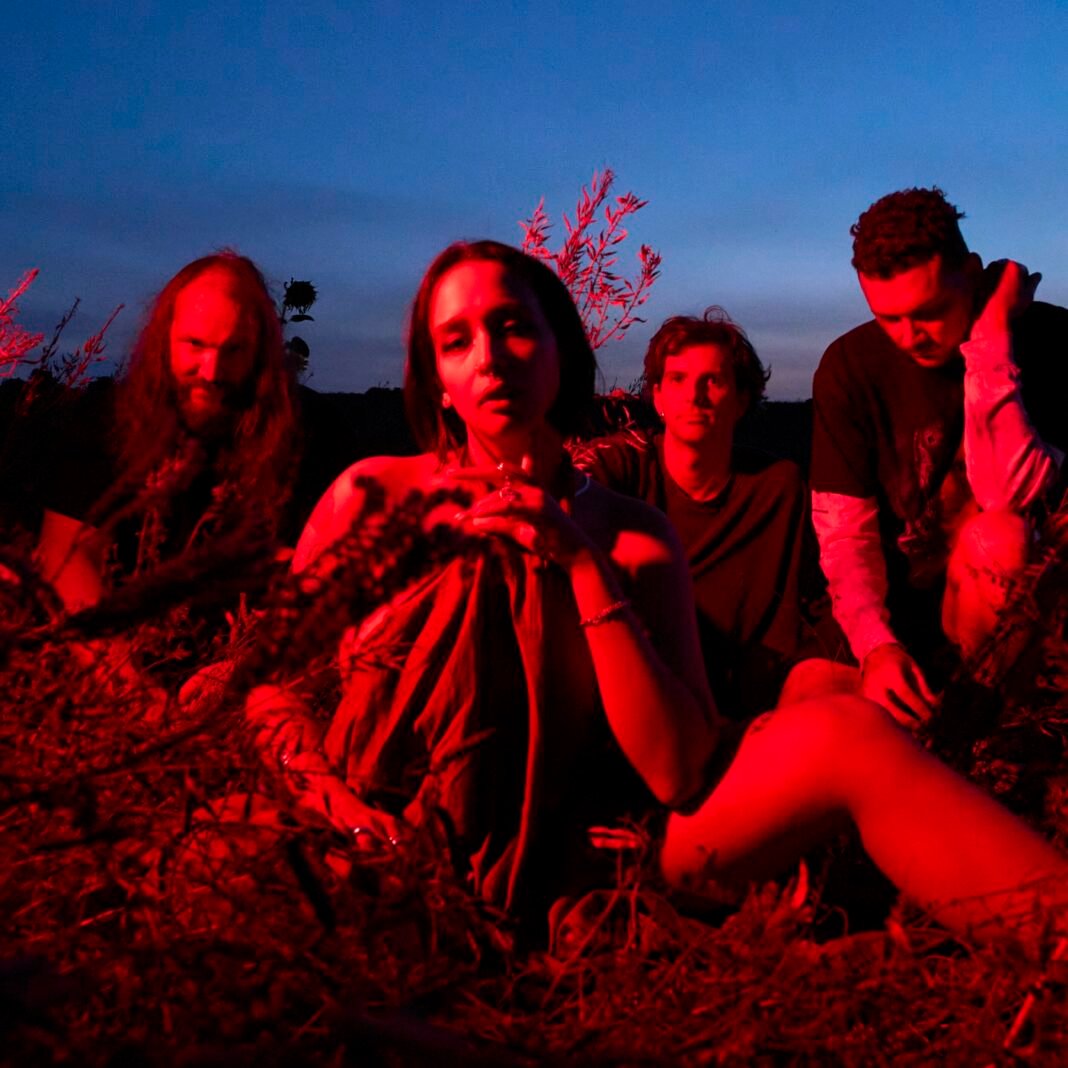 Four members of Mandy Indiana sit outdoors at dusk, lit by dramatic red light against a deep blue sky, with vocalist Valentine Caulfield centered in the foreground.