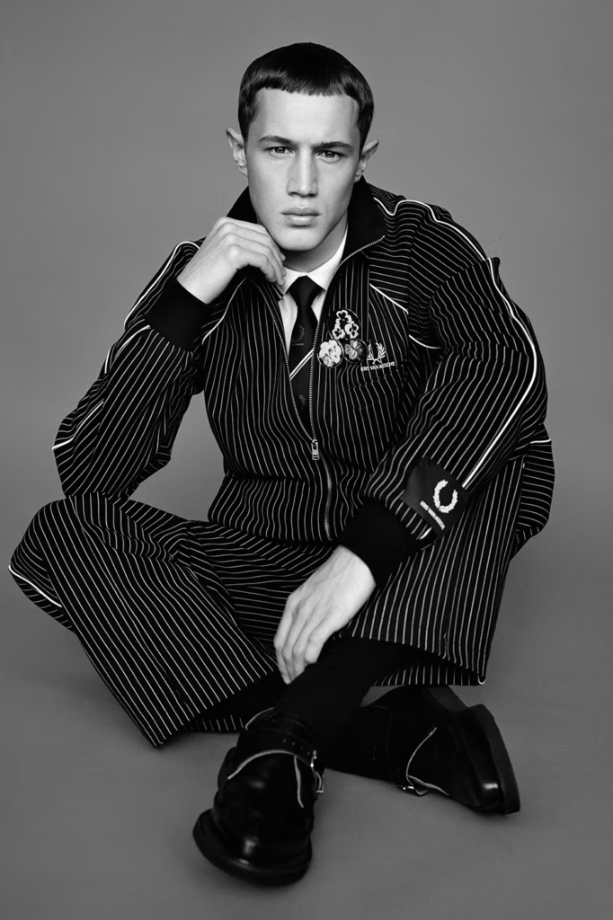 Black and white portrait of a male model wearing the Kris Van Assche x Fred Perry 2026 collaboration, featuring a striped tailored tracksuit with laurel wreath logo and decorative patches, styled in a modern uniform-inspired aesthetic.