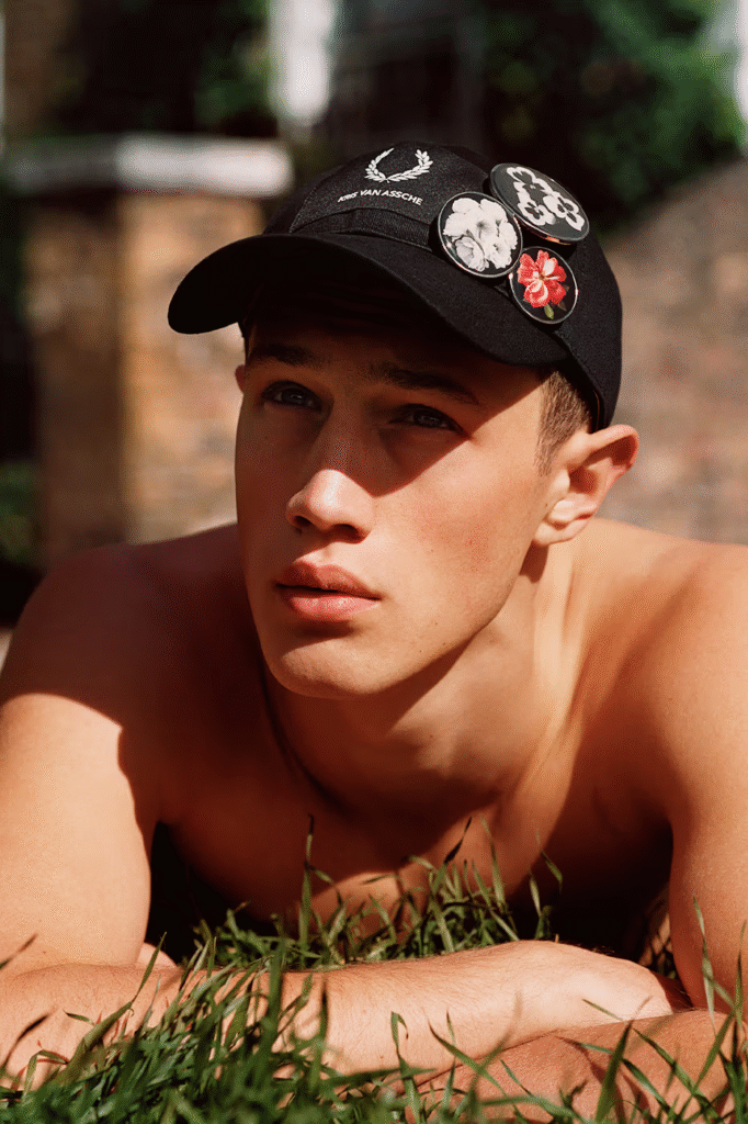 Close-up outdoor portrait of a male model wearing a black Kris Van Assche x Fred Perry cap with laurel wreath logo and decorative floral badges, styled in warm natural sunlight against a blurred background.