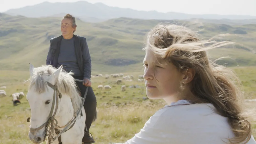 Two women in a mountainous grassland landscape, one riding a white horse while the other looks into the distance, with grazing sheep and rolling hills in the background.
