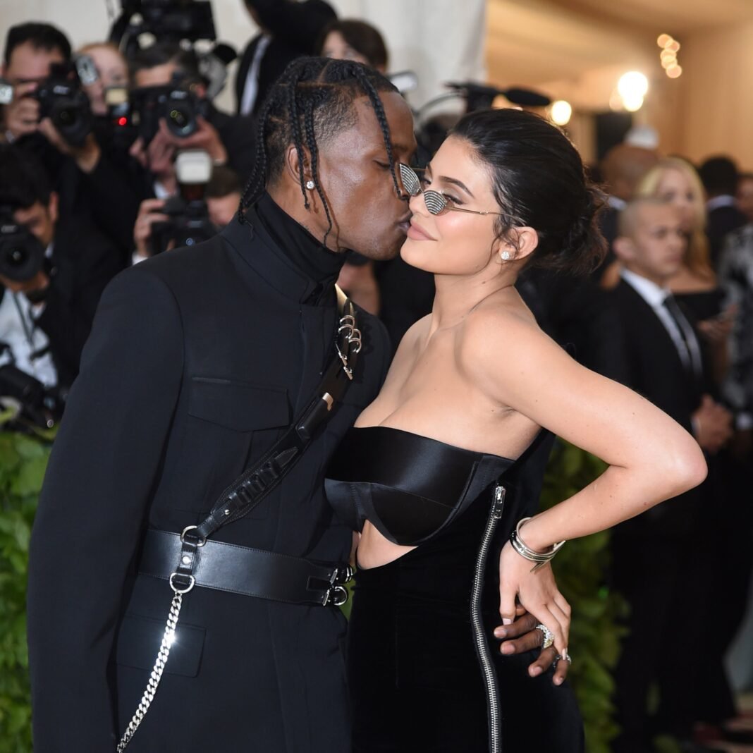 Travis Scott kisses Kylie Jenner on the cheek as they pose together on the red carpet, with Jenner wearing a black strapless gown and slim oval sunglasses, while photographers capture the moment in the background.