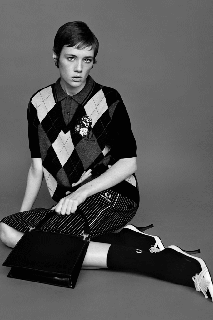 Black and white studio portrait of a model wearing the Kris Van Assche x Fred Perry 2026 collaboration, featuring an argyle knit top with decorative badges, striped skirt with laurel wreath logo, knee-high socks, and classic loafers in a modern uniform-inspired style.