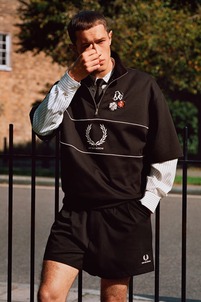 Outdoor fashion image of a male model wearing the Kris Van Assche x Fred Perry 2026 collaboration, featuring a black short-sleeve top with laurel wreath logo, layered striped shirt and tie, tailored shorts, and decorative badges in a modern uniform-inspired style.