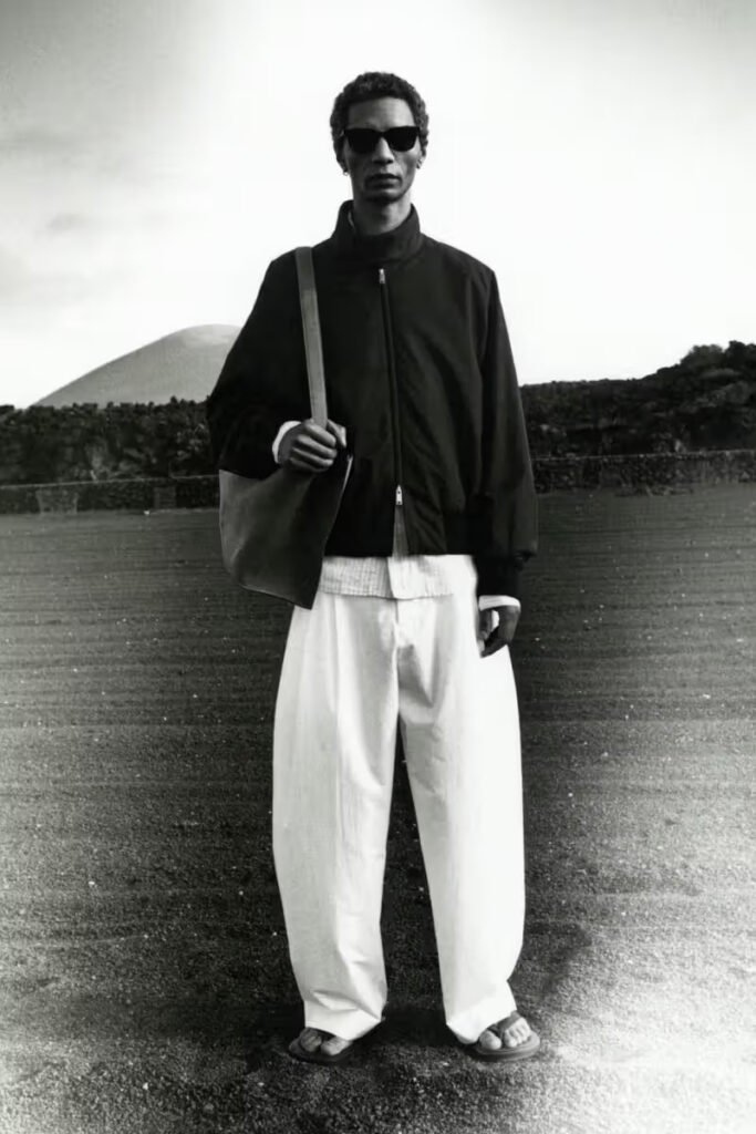 A-model-in-a-dark-zip-up-bomber-jacket-voluminous-white-wide-leg-trousers-sandals-and-sunglasses-carrying-a-leather-tote-standing-against-a-volcanic-landscape