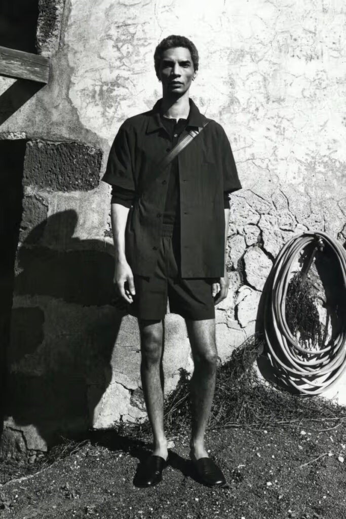 A-model-in-a-matching-dark-short-sleeve-shirt-and-shorts-set-crossbody-bag-and-loafers-standing-against-a-weathered-stone-wall