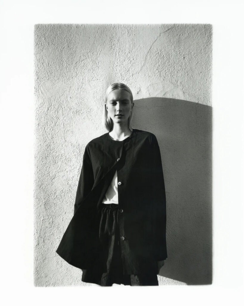 A-model-in-an-oversized-collarless-dark-jacket-and-wide-leg-shorts-shot-in-black-and-white-against-a-textured-wall-with-dramatic-shadow.