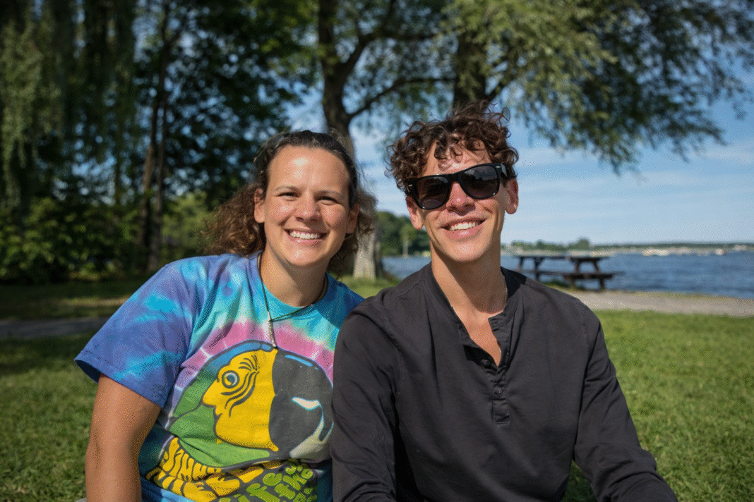 A woman in a tie-dye t-shirt and Matthew Brian Robertson in a black shirt and sunglasses smiling together at a sunny lakeside park.