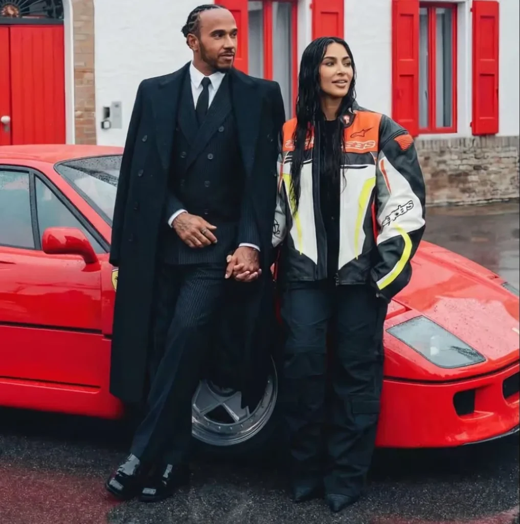 Lewis Hamilton and Kim Kardashian holding hands in front of a classic red Ferrari F40, he in a long black double-breasted coat, she in an Alpinestars racing suit.