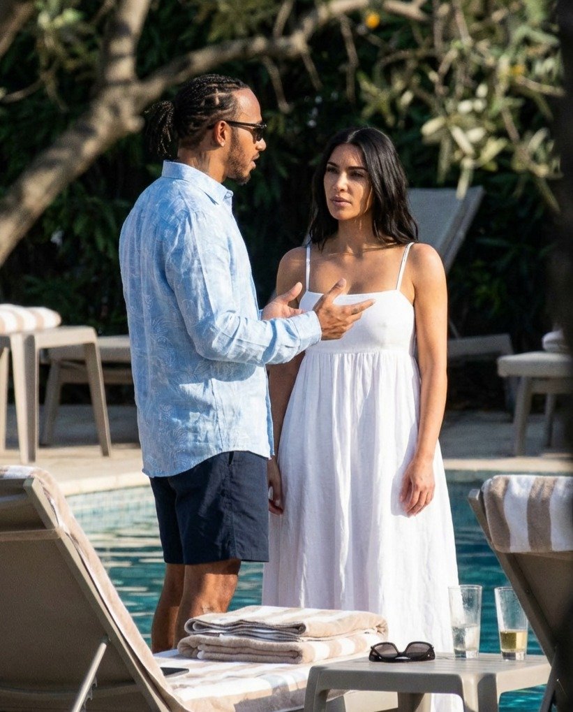 Lewis Hamilton and Kim Kardashian in conversation beside a sun-drenched pool, he in a light blue linen shirt and shorts, she in a flowing white sundress.