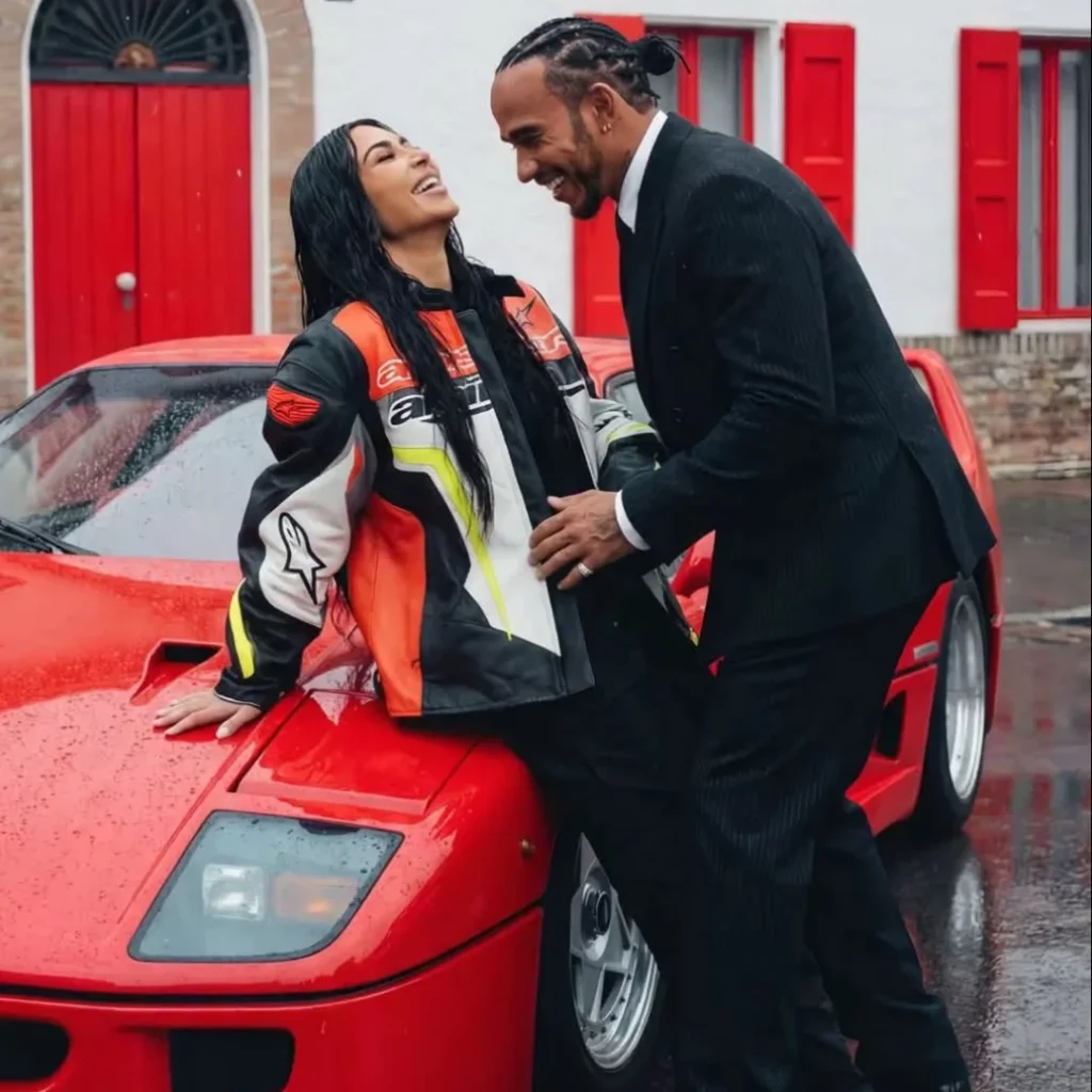 Lewis Hamilton and Kim Kardashian laughing together beside a classic red Ferrari F40 in the rain, she in a racing jacket, he in a black suit.