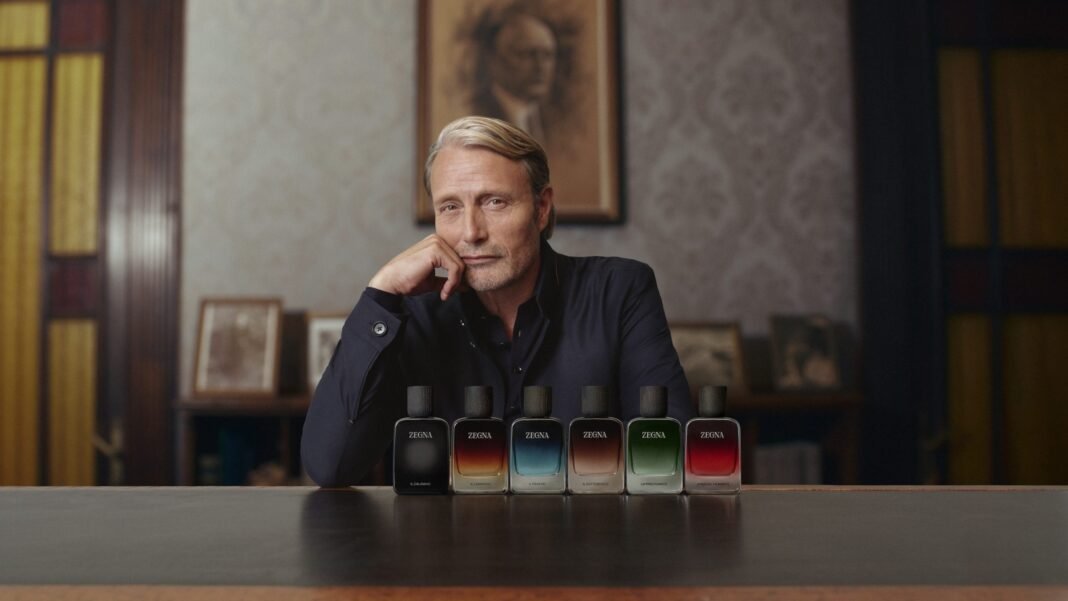 Mads Mikkelsen poses with the six ZEGNA MEMORIE fragrance bottles displayed on a table in a wood-paneled study
