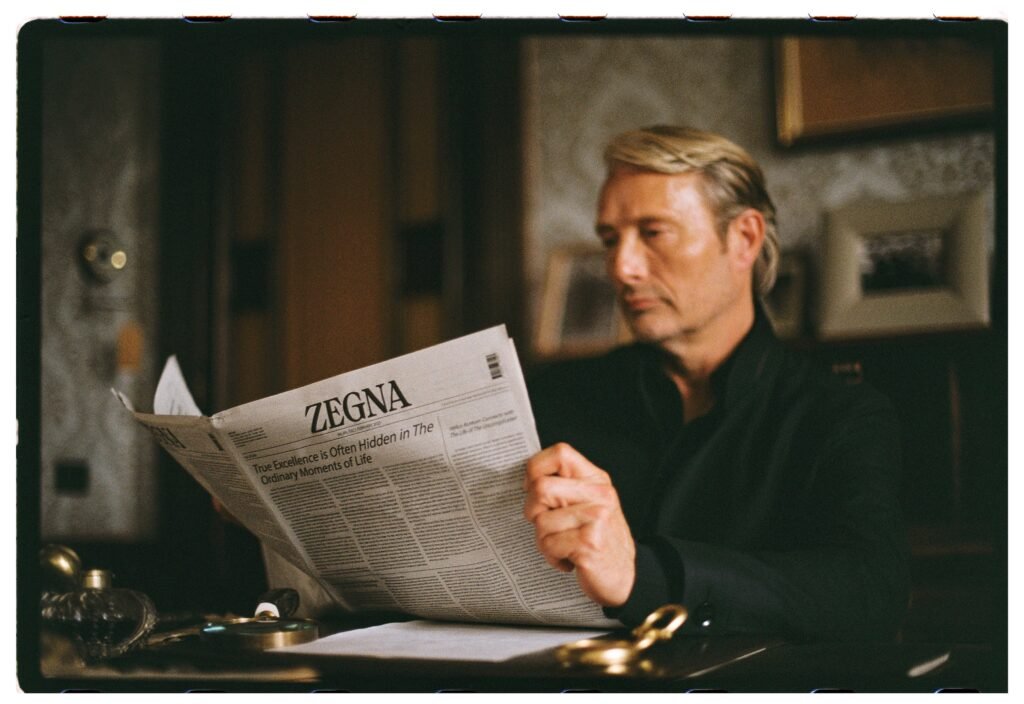 Mads Mikkelsen reads a ZEGNA branded newspaper at a vintage desk in a film still from the ZEGNA MEMORIE fragrance campaign