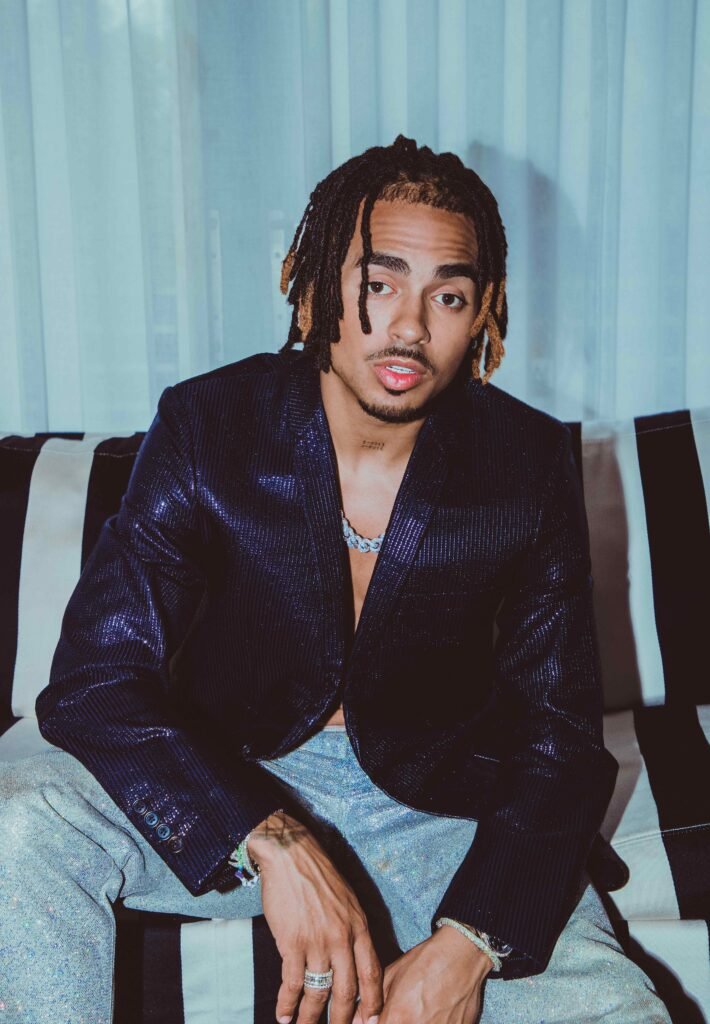 Ozuna sits on a striped chair wearing an open navy sequin blazer with iridescent glitter trousers and a blue Cuban link chain