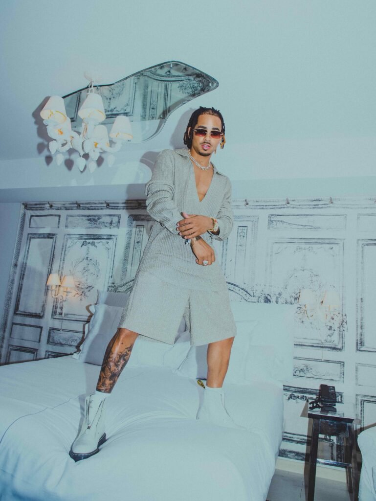 Ozuna stands on a white bed wearing a matching beige knit cardigan and shorts set with white combat boots and gold jewelry in a Flaunt Magazine editorial shoot