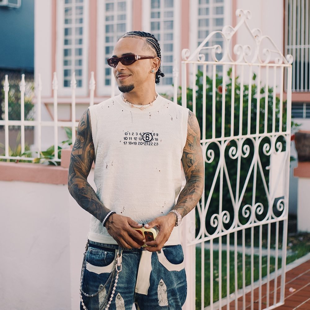 Ozuna stands outside a colorful Puerto Rico building wearing an MM6 Maison Margiela sleeveless top and tattooed arms on display