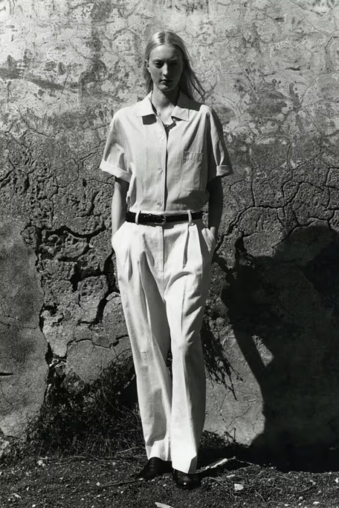 Studio-Nicholson-SS26-female-model-in-a-linen-short-sleeve-shirt-and-wide-leg-trousers-photographed-in-black-and-white-against-a-textured-stone-wall