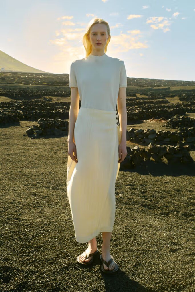 Studio-Nicholson-SS26-female-model-in-a-white-short-sleeve-top-and-cream-maxi-skirt-standing-on-a-volcanic-landscape-at-golden-hour