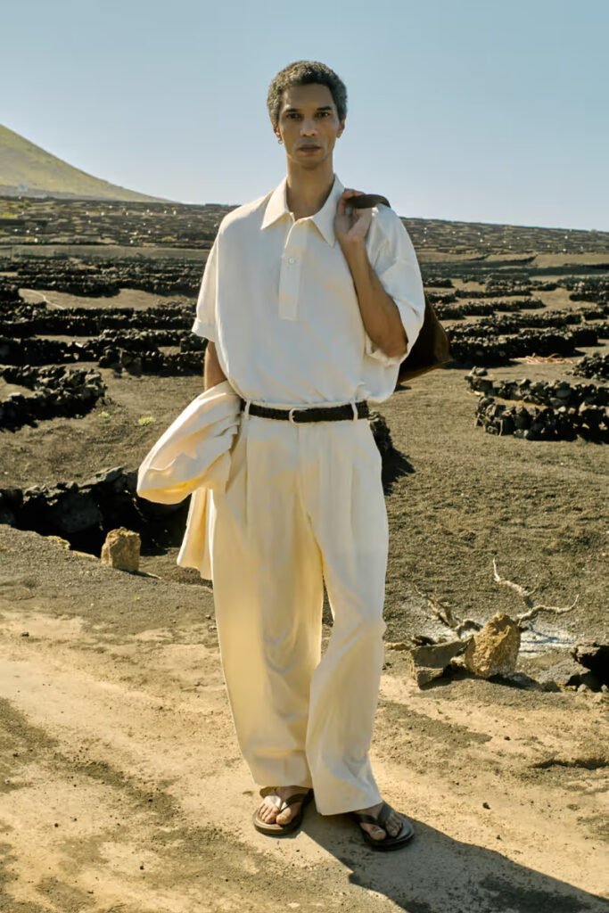 Studio-Nicholson-SS26-male-model-in-a-cream-polo-shirt-and-wide-leg-trousers-carrying-a-suede-jacket-on-a-volcanic-landscape