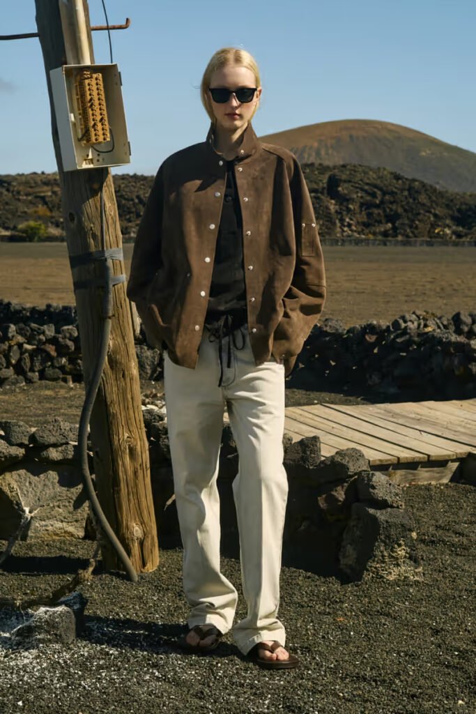 Studio-Nicholson-SS26-model-wearing-a-brown-suede-snap-button-jacket-and-cream-wide-leg-trousers-on-a-volcanic-outdoor-location