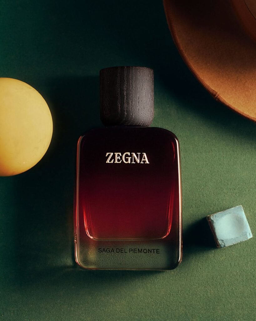 ZEGNA-MEMORIE-Saga-Del-Piemonte-fragrance-bottle-in-deep-crimson-gradient-styled-on-a-dark-green-surface-with-a-vintage-felt-hat-brim-and-chalk-block-as-props