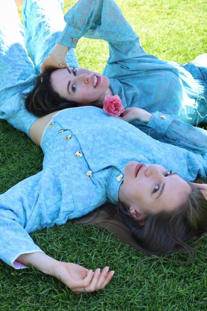 Two women lying on green grass wearing I Love Mirabelle blue floral quilted sets with gold heart buttons, one styled with a pink rose at the collar.