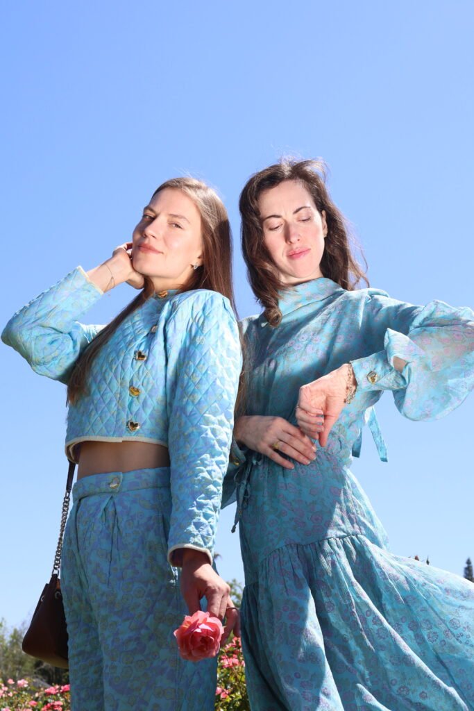 Two women wearing matching I Love Mirabelle sky-blue floral quilted and chiffon sets outdoors against a clear blue sky, one holding a pink rose.