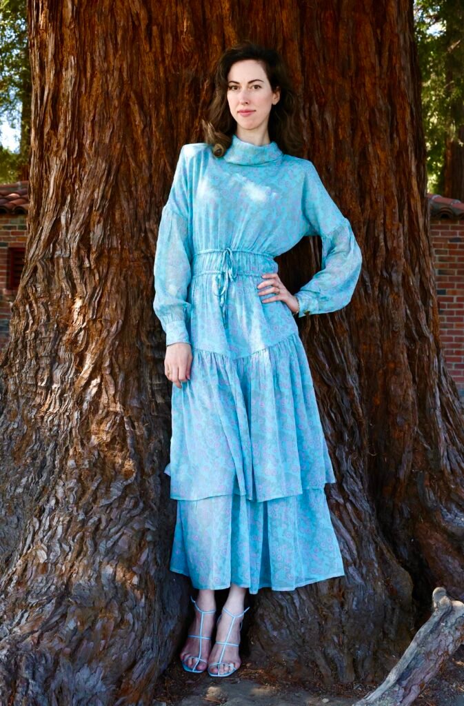 Woman standing against a redwood tree wearing an I Love Mirabelle sky-blue floral chiffon tiered maxi dress with drawstring waist and long sleeves, paired with strappy blue heels.