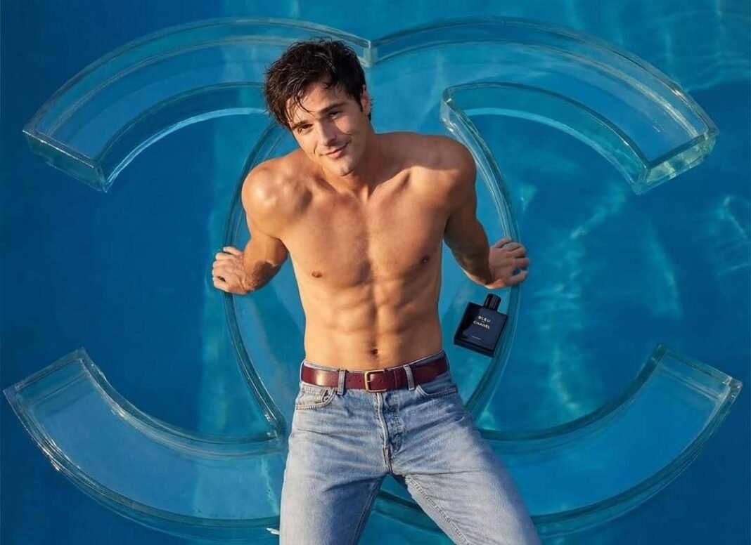 acob Elordi Bleu De Chanel 2026 campaign photo, shirtless holding fragrance bottle in front of oversized glass Chanel logo.