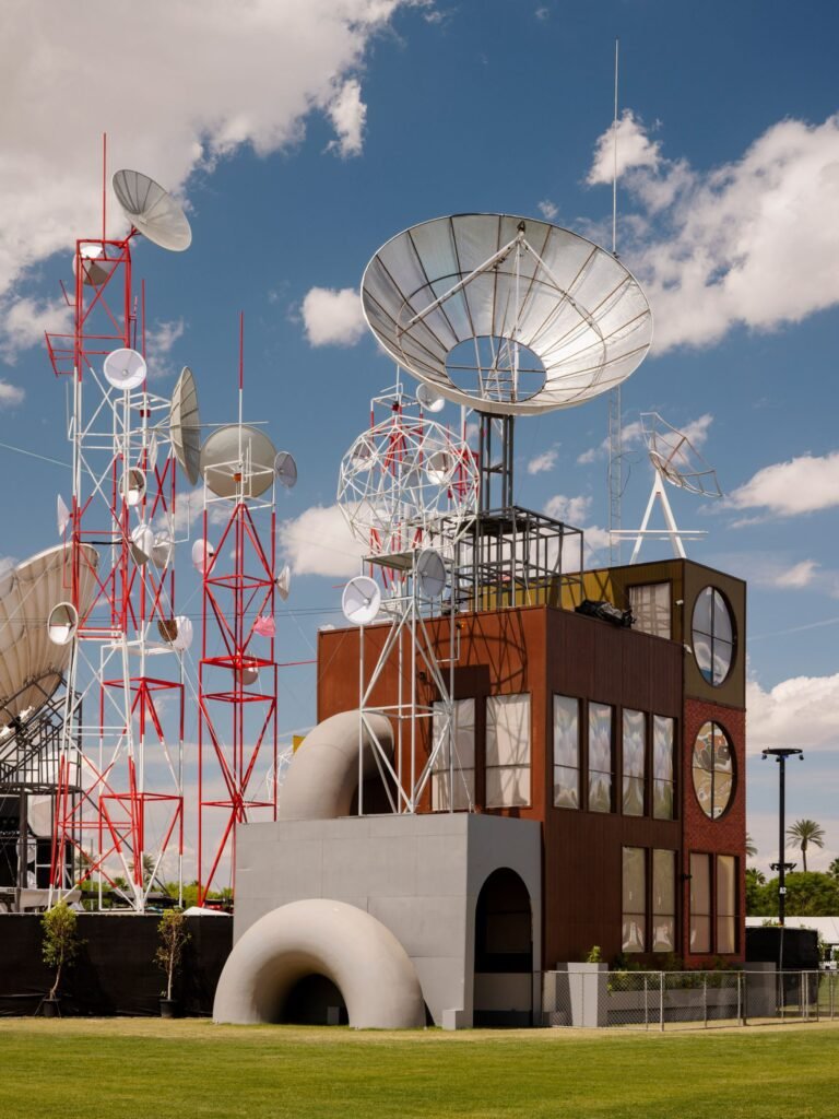 > Towering red and white satellite dish structures surround a brutalist building facade at Coachella 2026, set against a cloudy desert sky.
